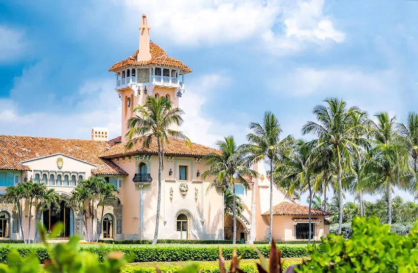 Mediterranean-style pink mansion surrounded by palm trees under a partly cloudy blue sky.